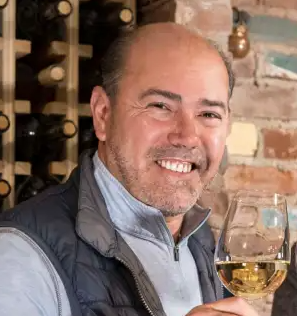 A smiling man holding a glass of white wine in a winery setting with wine bottles in the background.